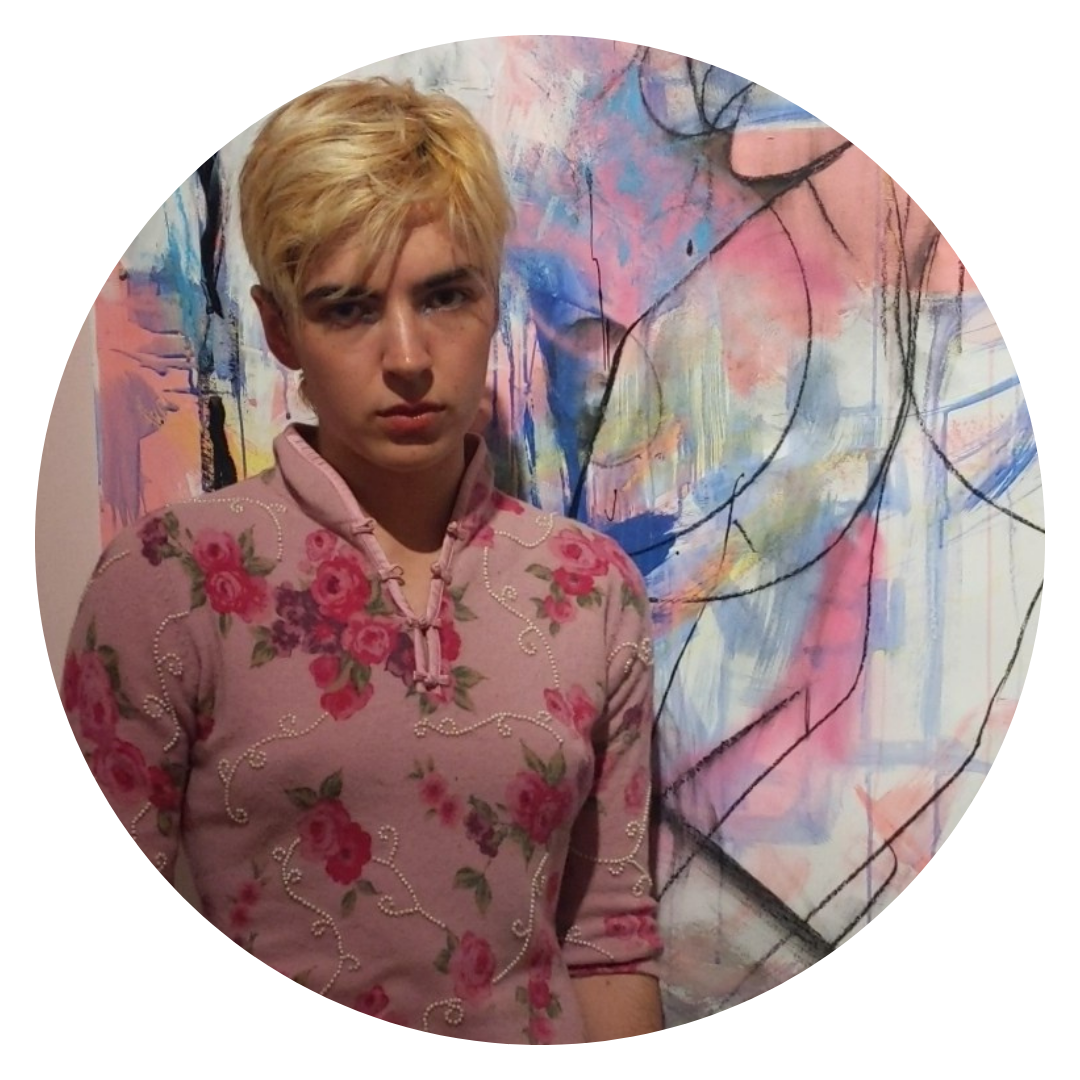 A photograph of the artist, Adam Charlton, standing in front of one of their paintings, which is abstract in tones of pastel pink, blue and charcoal. They are wearing a floral beaded top and have short blond hair.
