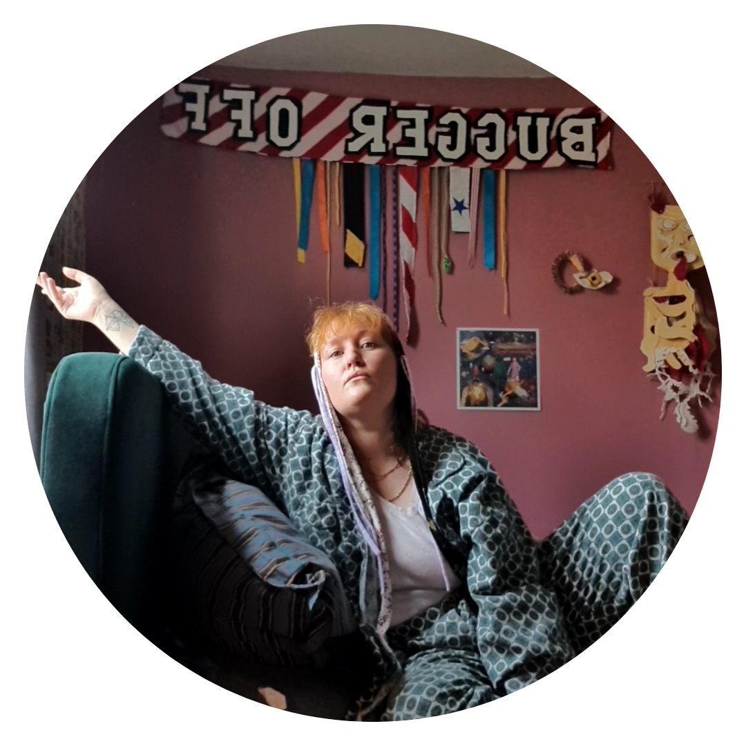 A photograph of the artist, Laura Lulika, sitting on a couch, an arm outstretched. In the background there is a scarf hanging up which says 'Bugger Off' in capital letters.