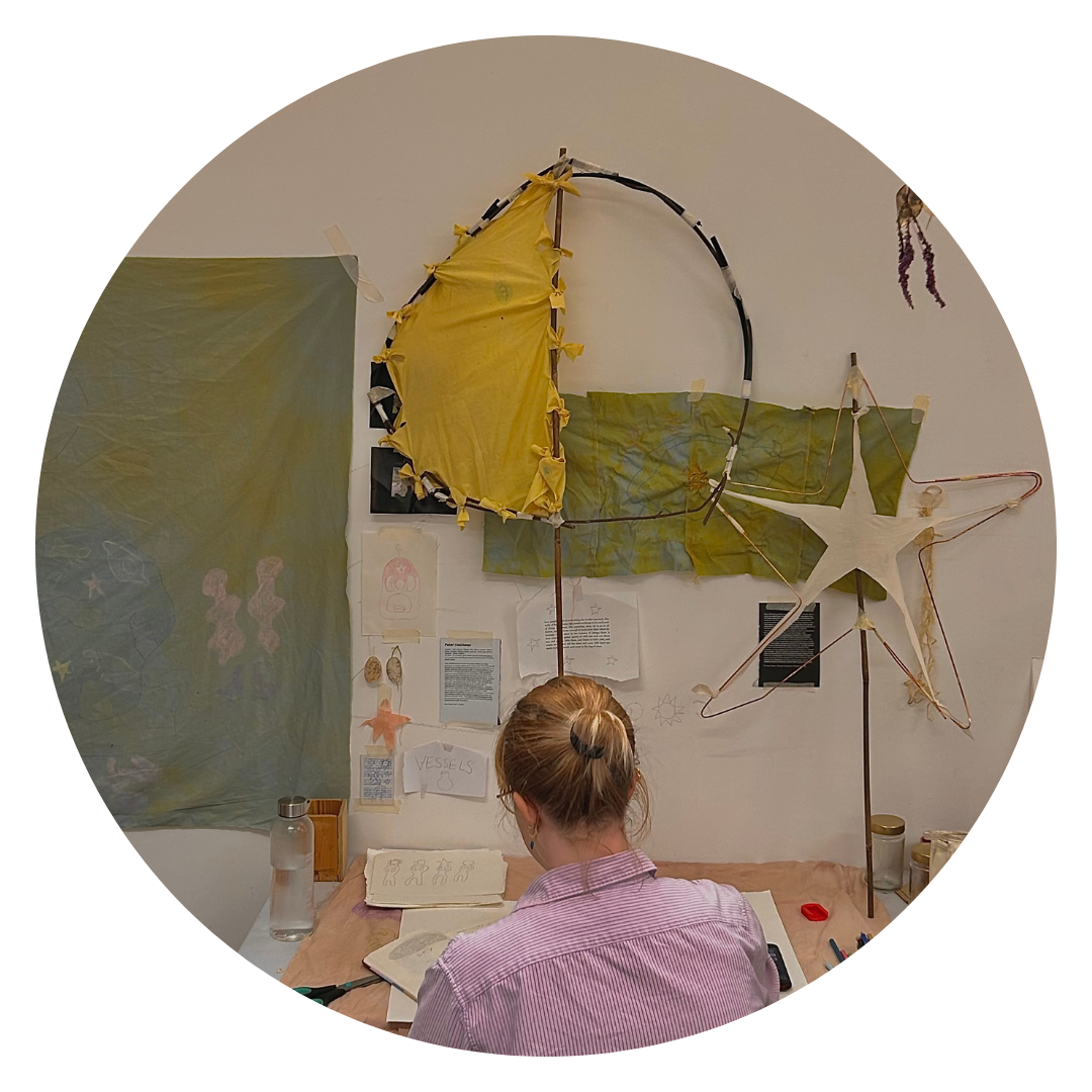 A photo of the artist Nancy Violet Downs, at work in their studio, taken from the back. Their hair is pulled back into a bun and they are wearing a striped pink shirt. On the wall are some works in progress made from fabric, wire and plant materials.