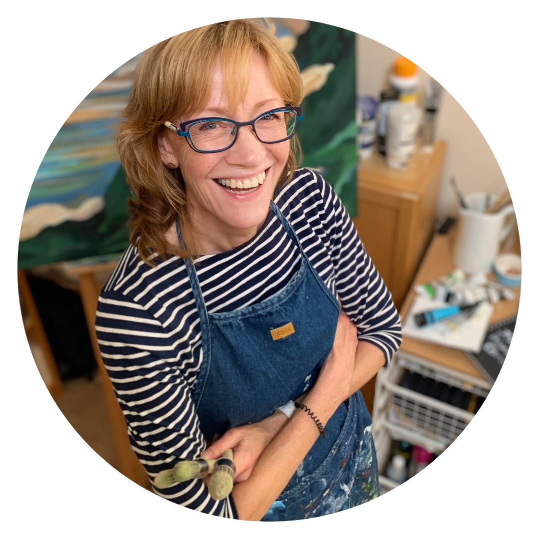 A photograph of the artist Liz Sergeant in their studio. They are smiling broadly and wearing glasses and a denim apron. In the background is a large painting in shades of green and blue.