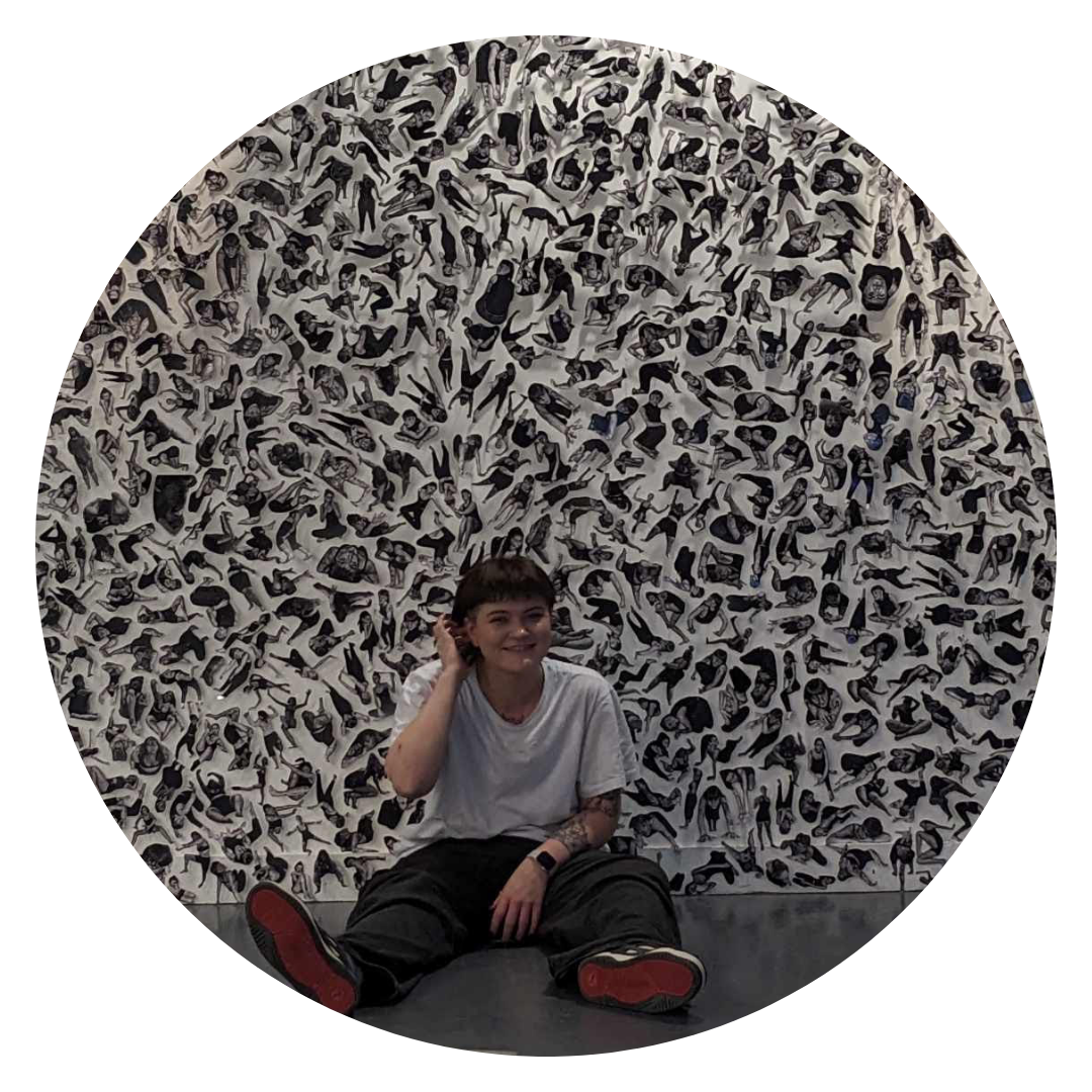 A photograph of the artist, Lee Oakley, sitting on the floor in front of their artwork, which features many black and white representations of female bodies. They are smiling and wearing trainers with red soles.